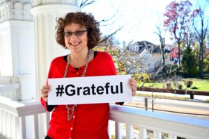 Head of School Naomi Reem stands on the porch of the South Campus holding a sign that reads "#Grateful".