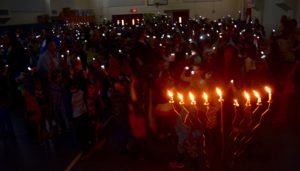 A full Chanukiah burns in the Gottesman Auditorium. The room is dark, dotted by small lights held by the students filling the gym.