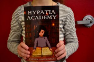 A student holds a copy of the book "Hypatia Academy" by Esther Goldenberg.