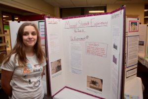 A sixth grader poses with her Inquiry Project board: "Magnet Strength Experiment"