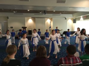 Students wearing blue and white costumes dance in a large social hall.
