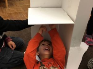 A boy lies on his back, affixing something to the underside of a half-built shelf. He is laughing as he works.