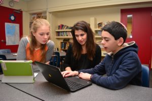 A sixth grade student shows Lisa Schopf something on his laptop as another student looks on.