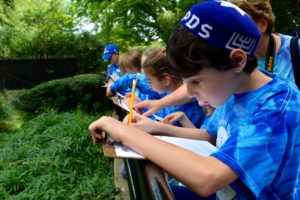 Students line up along the railing of an animal enclosure, balancing clipboards on the railing in order to take notes.