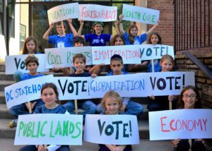 Students raise signs reading "Vote!" and naming the different issues the students studied, such as "Immigration" and "Globalization."