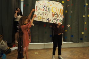 Two students hold up a sign on stage that reads, "Tikkun Olam Super Heroes to the Rescue!"