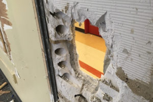 A hole in the wall of a classroom allows the viewer to see the hallway tile on the far side.