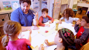 Kindergarten teacher Vas Pournaras sits with a group of students around a table.