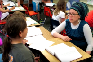 Judaic Studies teacher Zahava Bensimon sits with students, an open Chumash on the desk in front of them.