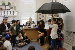 Children stand under an umbrella, acting out a Hebrew song in front of their parents at the Bayit Patuach (Open House).