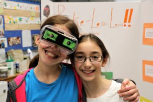 A fifth grader poses in front of her Invention Convention board while wearing her invention - a pair of modified sunglasses.