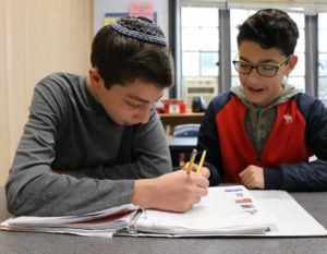 Two students sit together in a classroom, one of them speaking and the other writing in a notebook.