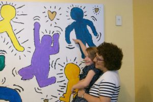 Head of School Naomi Reem lifts up a Pre-K student so she can point to her outline high up on the mural.