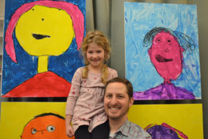 A Pre-Kindergarten student sits on her father's shoulder to pose next to her brightly colored "Happy" portrait at the Bayit Patuach (Open House).